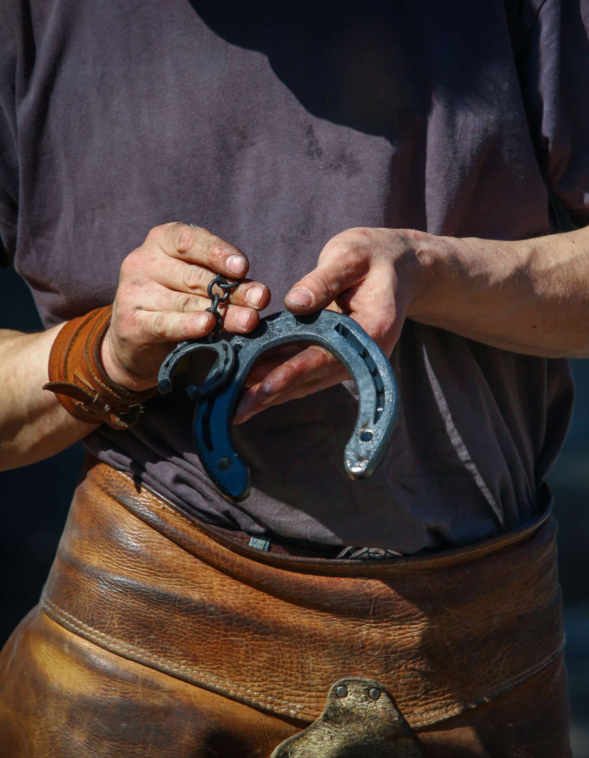 Mains d'un maréchal-ferrant avec un fer à cheval noir et une petite chaîne, portant un tablier en cuir marron.